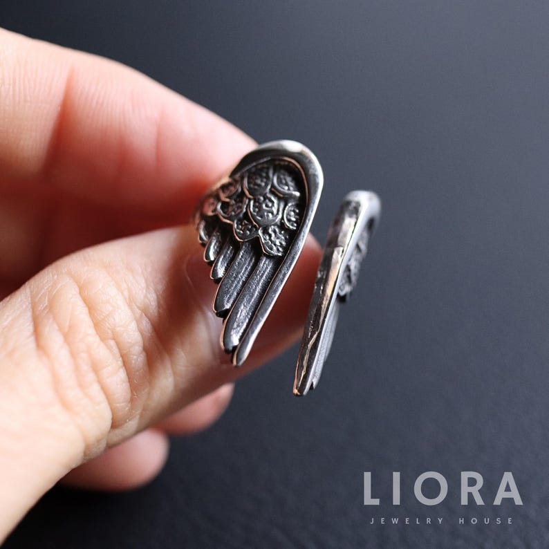 Puede incluir: Un anillo plateado con un dise&ntilde;o abierto, con un motivo de ala detallado. Las plumas del ala est&aacute;n intrincadamente talladas, creando una apariencia texturizada. El anillo se muestra en un dedo, destacando su naturaleza ajustable y la artesan&iacute;a de la joyer&iacute;a.
