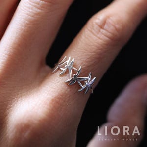 May include: A silver ring with a repeating dragonfly design. The ring is made of a shiny metal and has a delicate, openwork pattern. The dragonfly motifs are evenly spaced around the band.