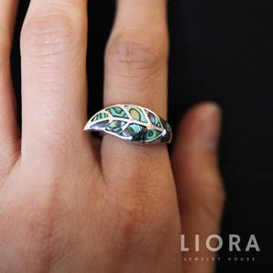 May include: A silver ring with an abalone inlay. The ring features a leaf-like design with iridescent green, blue, and silver tones. The ring is on a finger, with a black background.