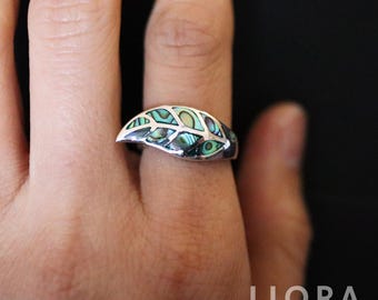 May include: A silver ring with an abalone inlay. The ring features a leaf-like design with iridescent green, blue, and silver tones. The ring is on a finger, with a black background.