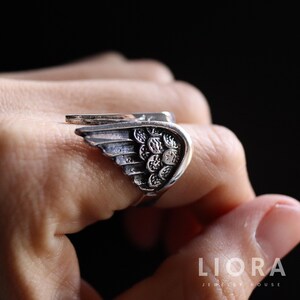 Puede incluir: Anillo plateado con dise&ntilde;o de ala. El anillo presenta detalles de plumas y una superficie texturizada. El ala envuelve el dedo, creando un accesorio &uacute;nico y elegante. El anillo se lleva en una mano sobre un fondo oscuro.