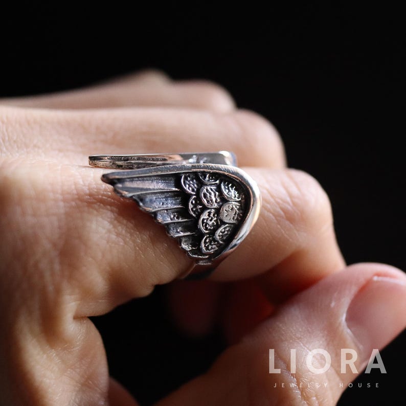Puede incluir: Anillo plateado con dise&ntilde;o de ala. El anillo presenta detalles de plumas y una superficie texturizada. El ala envuelve el dedo, creando un accesorio &uacute;nico y elegante. El anillo se lleva en una mano sobre un fondo oscuro.