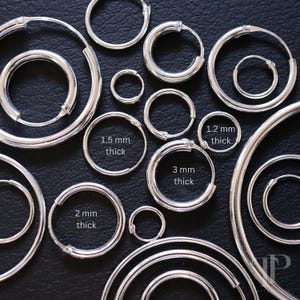Puede incluir: Una variedad de pendientes de aro de plata en diferentes tamaños y grosores. Los pendientes están dispuestos sobre una superficie negra. El texto de la imagen dice "1,2 mm de grosor", "1,5 mm de grosor", "2 mm de grosor", "3 mm de grosor".