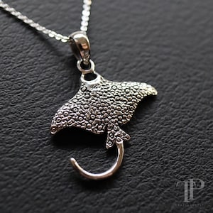 Puede incluir: Un collar con colgante plateado que presenta un diseño detallado de una manta raya. El colgante tiene una superficie texturizada y está suspendido de una delicada cadena. La raya tiene un cuerpo ancho y plano y una cola curvada.