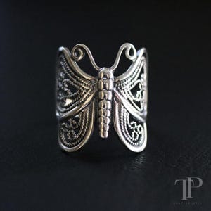 May include: A silver butterfly ring with an openwork design. The ring showcases detailed patterns on the butterfly's wings, body, and antennae. The ring is displayed against a dark backdrop, emphasizing its intricate design and craftsmanship.