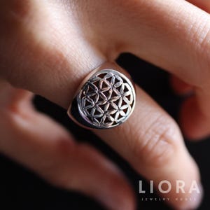 Op de afbeelding: Een zilveren ring met een gedetailleerd Flower of Life-ontwerp. De ring heeft een afgeronde voorkant met een opengewerkt patroon, dat het geometrische symbool laat zien. De merknaam "LIORA JEWELRY HOUSE" is zichtbaar.