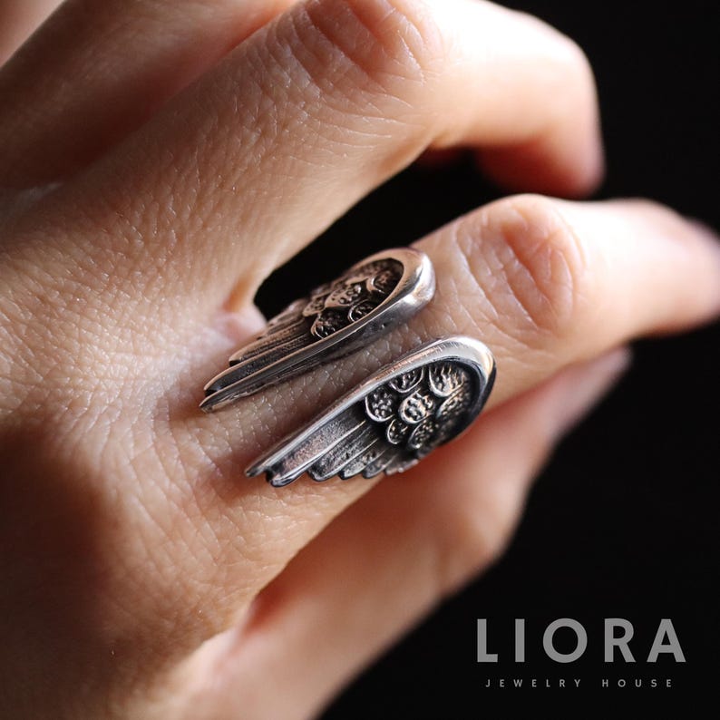 Puede incluir: Un anillo plateado con un dise&ntilde;o detallado de alas. El anillo envuelve el dedo, con cada ala extendi&eacute;ndose hacia afuera. La joya se encuentra sobre un fondo oscuro, resaltando los intrincados detalles de las alas.