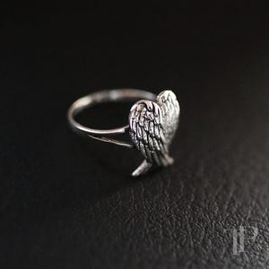 May include: A silver ring featuring a detailed wing design. The ring has a polished finish and is set against a dark background. The wing is intricately detailed, showcasing individual feathers.