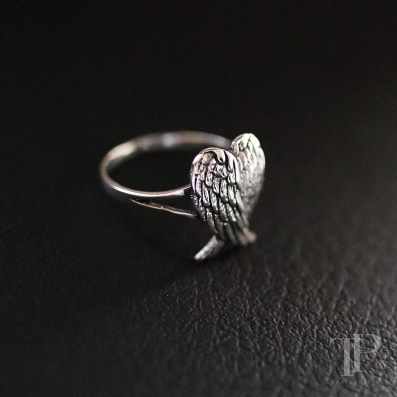 May include: A silver ring featuring a detailed wing design. The ring has a polished finish and is set against a dark background. The wing is intricately detailed, showcasing individual feathers.