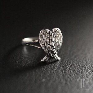 May include: A silver ring featuring a heart-shaped design of angel wings. The ring has a detailed feather texture and is set against a dark background. The ring is a piece of jewelry.