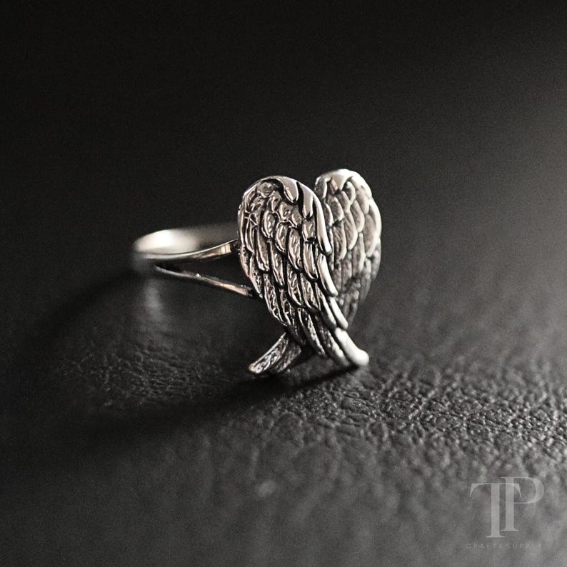 May include: A silver ring featuring a heart-shaped design of angel wings. The ring has a detailed feather texture and is set against a dark background. The ring is a piece of jewelry.