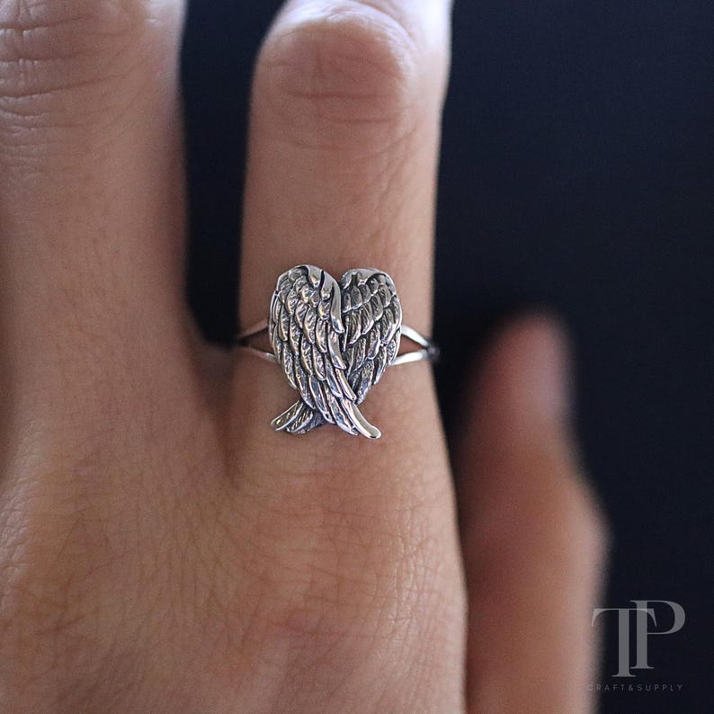 May include: A silver ring featuring a detailed angel wing design. The ring's focal point is the intricately carved wings, creating a heart shape. The ring is worn on a finger, showcasing the delicate craftsmanship and design.