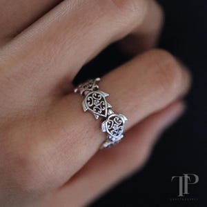 Puede incluir: Un anillo de plata con un diseño repetido de tortugas. Cada tortuga tiene un caparazón calado adornado con detalles decorativos. El anillo se muestra en un dedo, destacando el diseño intrincado y la artesanía.