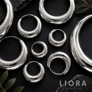 May include: A collection of polished silver-toned hoop earrings in various sizes, featuring a thick, rounded, and reflective surface. The earrings are arranged in a cluster, showcasing their smooth, metallic finish and circular design.