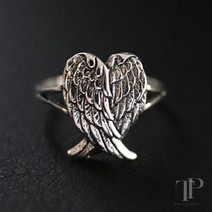 Op de afbeelding: Zilveren ring met gedetailleerd engelenvleugelontwerp. De vleugels kruisen elkaar aan de basis en vormen een hart. De ingewikkelde details van de ring worden benadrukt tegen een donkere achtergrond.