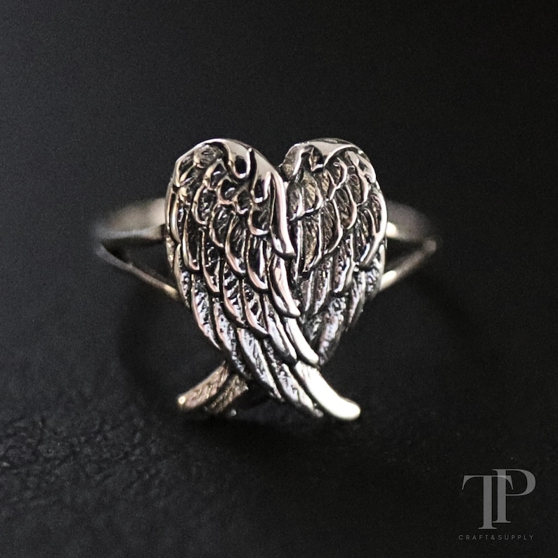 May include: A silver ring featuring a detailed design of angel wings. The wings are crossed at the base, forming a heart shape. The ring is set against a dark background, highlighting the intricate details of the wings.