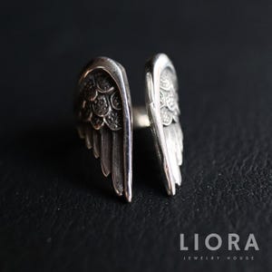 Puede incluir: Un anillo plateado con un dise&ntilde;o abierto, con dos formas de alas detalladas. Las alas tienen detalles de plumas texturizadas y est&aacute;n dise&ntilde;adas para envolver un dedo. El anillo se muestra sobre un fondo oscuro.