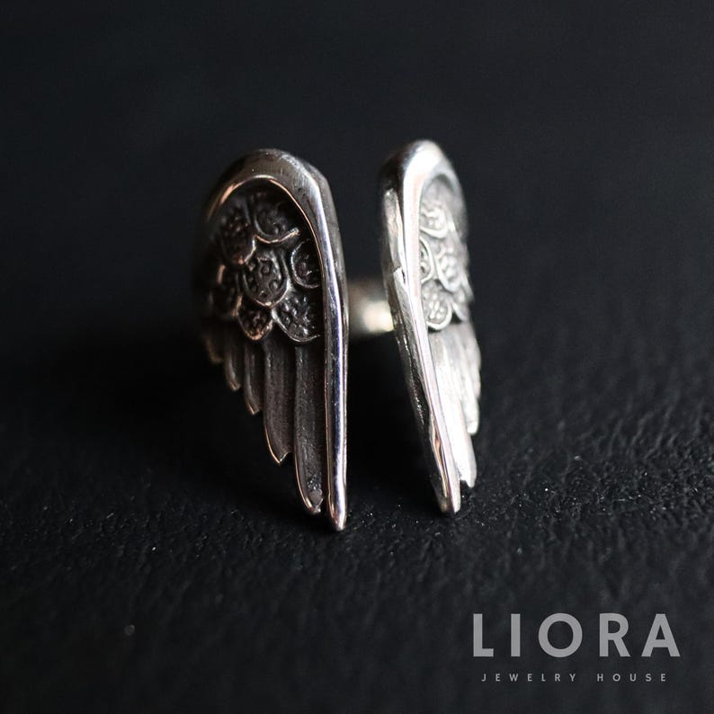 Puede incluir: Un anillo plateado con un dise&ntilde;o abierto, con dos formas de alas detalladas. Las alas tienen detalles de plumas texturizadas y est&aacute;n dise&ntilde;adas para envolver un dedo. El anillo se muestra sobre un fondo oscuro.