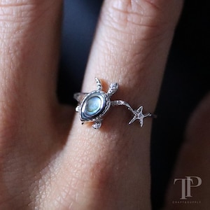 May include: A silver ring with a sea turtle design, featuring a pearlescent shell and a starfish. The ring is worn on a finger, highlighting the detailed craftsmanship. This ocean-themed ring is a delicate piece of jewellery.