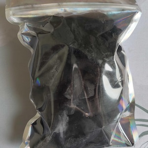 Puede incluir: Una bolsa de plástico transparente llena de objetos oscuros de forma irregular. La bolsa tiene una parte superior plateada e iridiscente con un pequeño agujero para colgar. El contenido parece ser carbón vegetal o un material similar.