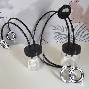 Può includere: Due barattoli di vetro trasparente con coperchi neri e bracci in metallo nero curvi. Ogni barattolo ha un disegno a corda bianca all'interno. Due anelli decorativi in bianco e nero sono davanti ai barattoli. Lo sfondo ha un motivo floreale.