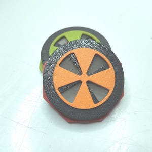 Könnte beinhalten: Ein Stapel runder, mehrfarbiger Kunststofffilter mit einem strukturierten schwarzen Außenring und einem zentralen sternförmigen Einsatz in Orange oder Limettengrün. Jeder Filter hat ein feines Metallgitter, das durch die Ausschnitte sichtbar ist.