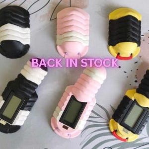 May include: Six segmented, pill-bug-shaped digital counters in black and white, pink and white, and yellow and black colour combinations. Three counters feature a rectangular digital display screen. The text BACK IN STOCK is centred in the image.