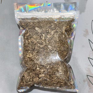 Puede incluir: Una bolsa de plástico transparente, holográfica y resellable, llena de una cantidad de corteza de madera triturada o material botánico seco de color marrón claro. La bolsa presenta un acabado metálico iridiscente en los bordes.