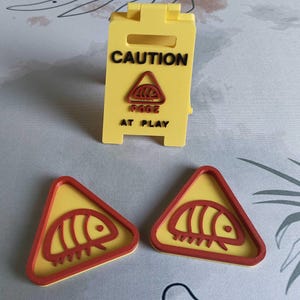 Op de afbeelding: Geel plastic bord met het woord "CAUTION" in zwart. Het bord heeft een rode driehoek met een gestileerde insectengrafiek en de woorden "RODE AT PLAY". Er zijn ook twee bijpassende rode en gele waarschuwingsborden met insectenthema's aanwezig.