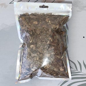 Può includere: Un sacchetto trasparente e richiudibile pieno di foglie marroni essiccate. La borsa ha una parte superiore argentata e iridescente con un piccolo foro per appenderla. Le foglie sono piccole e di forma irregolare, con varie tonalità di marrone.