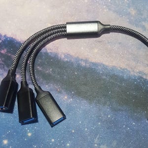 Könnte beinhalten: Ein schwarzer USB-Hub mit drei USB-Anschlüssen und einem einzelnen USB-Anschluss. Das Kabel ist mit einem schwarz-grauen Muster geflochten. Der Hub dient zum Anschluss mehrerer Geräte an einen einzelnen USB-Anschluss.