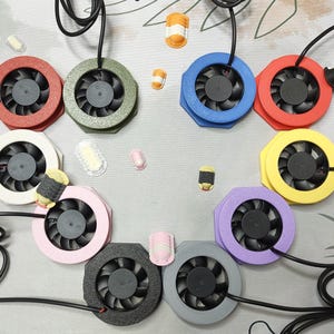 Pode incluir: Uma coleção de dez pequenas ventoinhas de arrefecimento circulares com pás pretas e molduras em várias cores, incluindo vermelho, verde-azeitona, azul, branco, amarelo, rosa, roxo, preto e cinzento, cada uma com um cabo de alimentação preto.