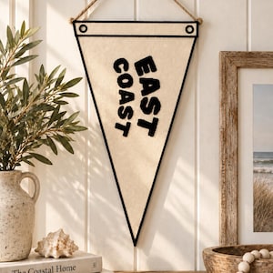 May include: A triangular cream-colored felt pennant featuring the black text "EAST COAST" oriented vertically. The pennant is framed by a black border, includes two black grommets at the top corners, and hangs from a brown rope attached to a wall.