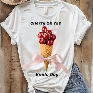 May include: White t-shirt with the text "Cherry On Top Kinda Day" and a graphic of cherries in an ice cream cone with a pink bow. The shirt is paired with denim shorts. A straw hat and sunglasses are also visible.