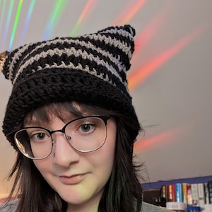 May include: A person wearing a black and gray striped crocheted cat ear hat with a silver chain necklace and a round pendant.