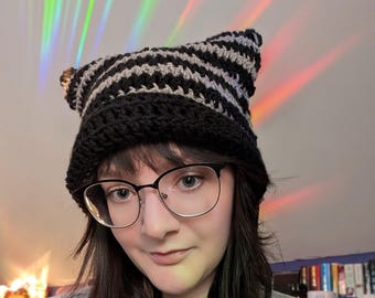 Peut inclure: Une personne portant un bonnet en crochet à oreilles de chat rayé noir et gris avec un collier en chaîne argentée et un pendentif rond.