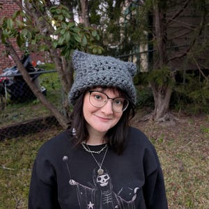 May include: A person wearing a grey crocheted cat hat with ears, black framed glasses, and a black shirt with a skeleton graphic.