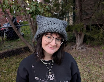 以下が含まれることがあります： 猫の耳が付いたグレーのクロシェハット、黒縁メガネ、骸骨のグラフィックがプリントされた黒いシャツを着た人。