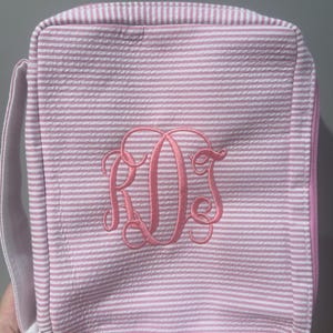May include: Pink and white striped seersucker bag with a pink zipper and handle. The bag features a pink monogram of the letters R, D, and S. The bag is rectangular and has a seersucker texture.