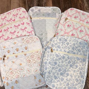 May include: Five rectangular quilted fabric cases with gold-tone zippers. The designs include pink bows, blue and white floral patterns, and patchwork motifs in soft pastel colors. Each case features a front zippered pocket and a handle.