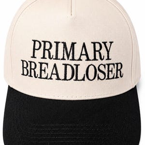May include: A two-tone baseball cap featuring a cream-colored crown and a black brim. The front of the crown displays the embroidered black text "PRIMARY BREADLOSER" in a serif font. The cap has a structured five-panel design with a black button.