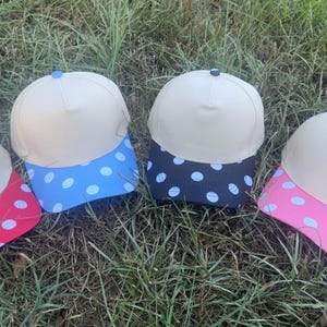 May include: Four baseball caps with cream-colored crowns and brims featuring white polka dots. The brims are red, blue, black, and pink respectively. Each cap has a small button on the top center matching the color of its respective brim.