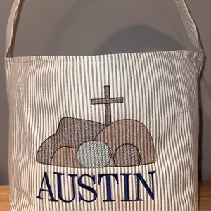 May include: A striped fabric Easter basket with a handle. The front features an embroidered design of a cross above a tomb, with the name "AUSTIN" embroidered below in navy blue.