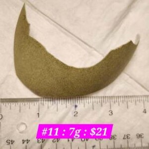 May include: A tan-coloured, curved object with a textured surface, possibly a piece of pottery or a similar craft item. A ruler is placed below the object, with measurements in centimetres. The object is approximately 7.6 cm long. The text "#11: 7g : $21" is visible.