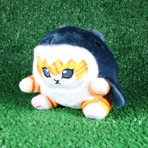 May include: A plush toy shaped like a shark with a white, orange, and black striped face. The toy has large, black eyes and a small, pink nose. The shark is wearing a white, fluffy coat with a black, shark fin on top.