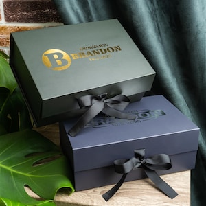 May include: Two gift boxes, one gray-green and one navy blue, are stacked. The gray-green box has a gold-colored initial and the name "Brandon" printed on it. Both boxes have a matching ribbon tied in a bow.