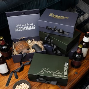 May include: Gift boxes in navy and green with black ribbon. One box is open, revealing a leather koozie, a small bottle, and a black tumbler. The open box has the text "Will you be my GROOMSMAN?" printed on the inside.