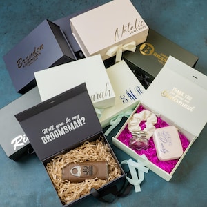 May include: An assortment of gift boxes in various colors, including navy, white, and olive green. Some boxes are open, revealing gifts like a cigar cutter, scrunchie, and small purse. Text on the boxes includes names and phrases like "Will you be my Groomsman?"