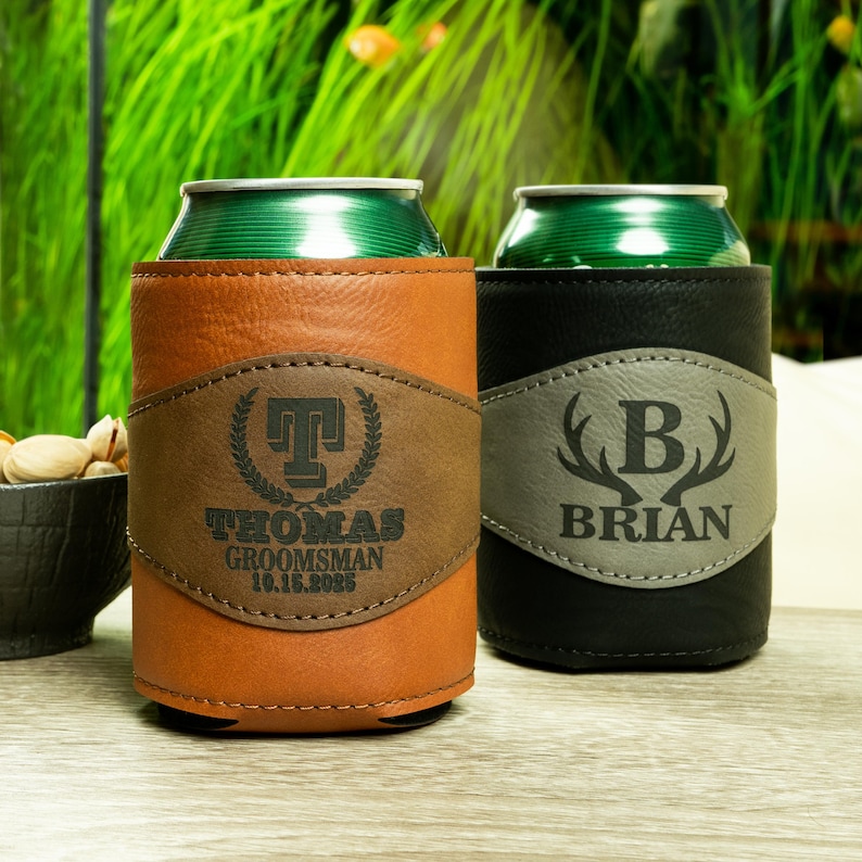 May include: Two personalized leather can coolers. The brown cooler has the name "Thomas" and the text "Groomsman 10.16.2023". The black cooler has the name "Brian" and a graphic of antlers with the letter "B" in the center.
