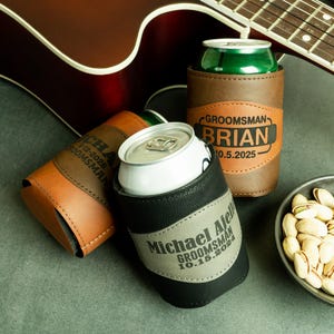 May include: Three personalized leather can coolers with brown, black, and gray leather. The coolers have the names "Brian", "Michael", and "Ajeli" with the text "Groomsman" and the date of the wedding.